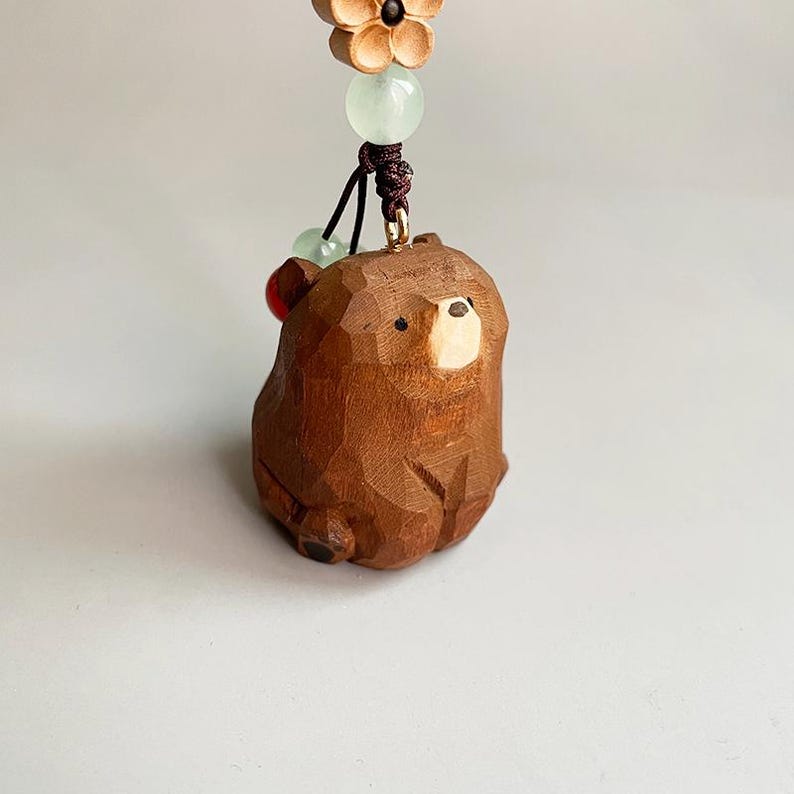 May include: A carved wooden bear figurine with a light brown finish. The bear is suspended by a dark brown cord, adorned with green and red beads, and a wooden flower. The bear's face has a lighter wood color.