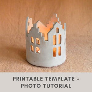 Könnte beinhalten: Ein weißer Kerzenhalter aus Keramik in Form einer Miniaturstadt mit ausgeschnittenen Fenstern und Dächern. Im Inneren scheint warmes Licht von einer brennenden Kerze. Der Text "PRINTABLE TEMPLATE + PHOTO TUTORIAL" befindet sich unten.