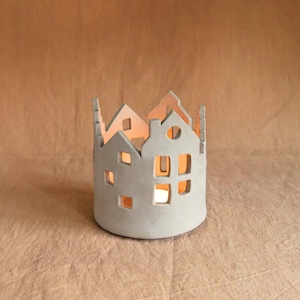 Pu&ograve; includere: Un portacandele in ceramica grigia a forma di fila di case. Le case hanno finestre e porte ritagliate, con un bagliore caldo che emana dall'interno. Un oggetto decorativo per l'arredamento della casa.