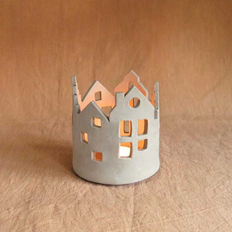 Pu&ograve; includere: Un portacandele in ceramica grigia a forma di fila di case. Le case hanno finestre e porte ritagliate, con un bagliore caldo che emana dall'interno. Un oggetto decorativo per l'arredamento della casa.