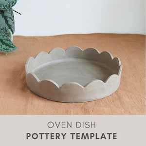 Op de afbeelding: Een ronde, lichtgrijze ovenschaal met een geschulpte rand. De schaal is gemaakt van een mat materiaal en staat op een bruinachtige ondergrond. De tekst "OVEN DISH POTTERY TEMPLATE" staat onderaan de afbeelding.