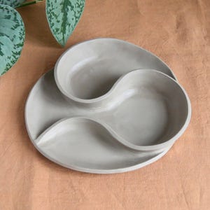 May include: A gray ceramic serving dish with a unique, flowing design. The dish features two connected, wave-like compartments, perfect for serving snacks or appetizers. The dish is set against a neutral background, highlighting its minimalist aesthetic.