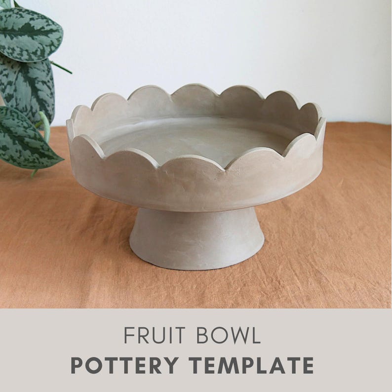 Op de afbeelding: Een beige aardewerken fruitschaal met een geschulpte rand, op een voetstuk. De kom staat op een bruin oppervlak. De tekst "FRUIT BOWL POTTERY TEMPLATE" staat onderaan de afbeelding.