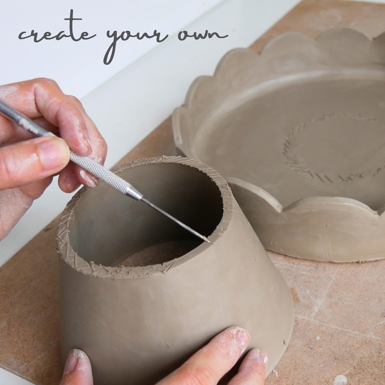 Op de afbeelding: Een persoon gebruikt een metalen gereedschap om een lichtbruine kleipot te vormen. De pot is cilindrisch met een uitlopende basis. Een bord met geschulpte rand staat op de achtergrond. De tekst "create your own" staat bovenaan.
