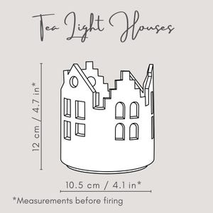 Pu&ograve; includere: Un portacandele in ceramica bianca a forma di fila di case. Presenta finestre rettangolari e ad arco e il testo "Tea Light Houses". Misura 12 cm di altezza e 10,5 cm di larghezza.
