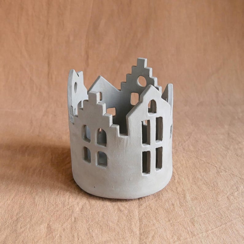 Pu&ograve; includere: Un portacandele cilindrico grigio con un design a forma di casa. Il supporto presenta finestre e tetti ritagliati, creando una silhouette di paesaggio urbano. Il pezzo in ceramica &egrave; di colore neutro, adatto a vari stili di arredamento.