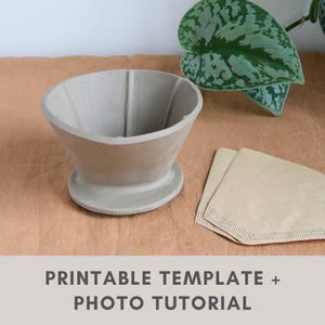 Puede incluir: Un vertedor de café de cerámica gris claro con un diseño acanalado, sobre una base pequeña y redonda. Dos filtros de café de papel marrón doblados están junto al vertedor. El texto "PRINTABLE TEMPLATE + PHOTO TUTORIAL" está en la parte inferior.