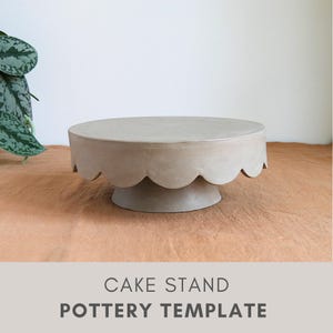 Op de afbeelding: Een neutraal gekleurde taartstandaard met een geschulpte rand. De standaard heeft een cirkelvormige bovenkant en een voetstuk. De woorden "CAKE STAND" en "POTTERY TEMPLATE" staan eronder gedrukt. De standaard is gemaakt van aardewerk.