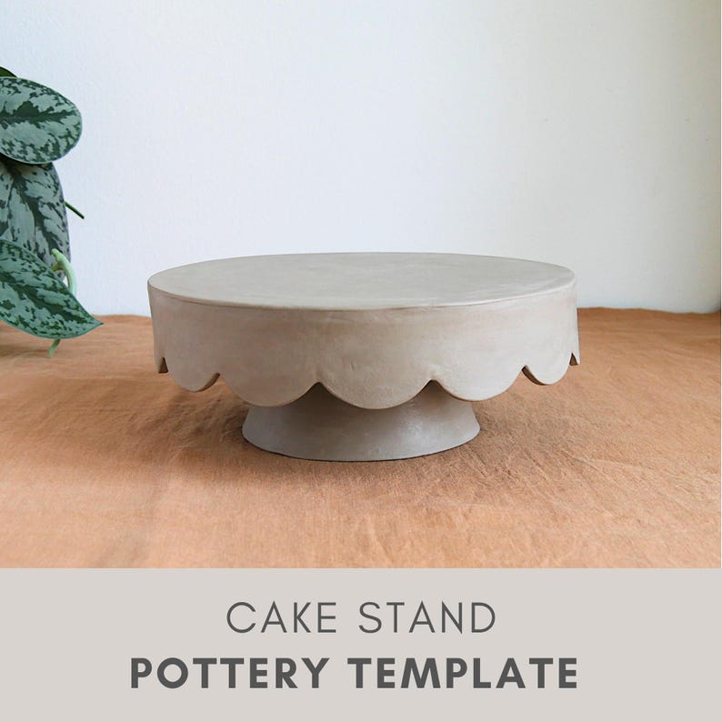 Op de afbeelding: Een neutraal gekleurde taartstandaard met een geschulpte rand. De standaard heeft een cirkelvormige bovenkant en een voetstuk. De woorden "CAKE STAND" en "POTTERY TEMPLATE" staan eronder gedrukt. De standaard is gemaakt van aardewerk.