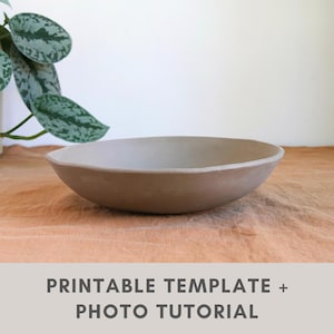 Peut inclure: Un bol en céramique marron clair fait à la main, de conception large et peu profonde. Le bol est posé sur une surface texturée de couleur beige. Le texte "PRINTABLE TEMPLATE + PHOTO TUTORIAL" est au bas de l'image. Une plante verte est en arrière-plan.