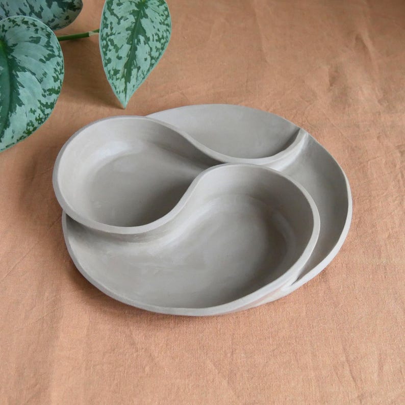 May include: A light gray ceramic serving dish with a unique, flowing design. The dish features two interconnected, curved sections and a circular outer rim. The neutral color and organic shape make it suitable for various uses, such as serving snacks or displaying small items.