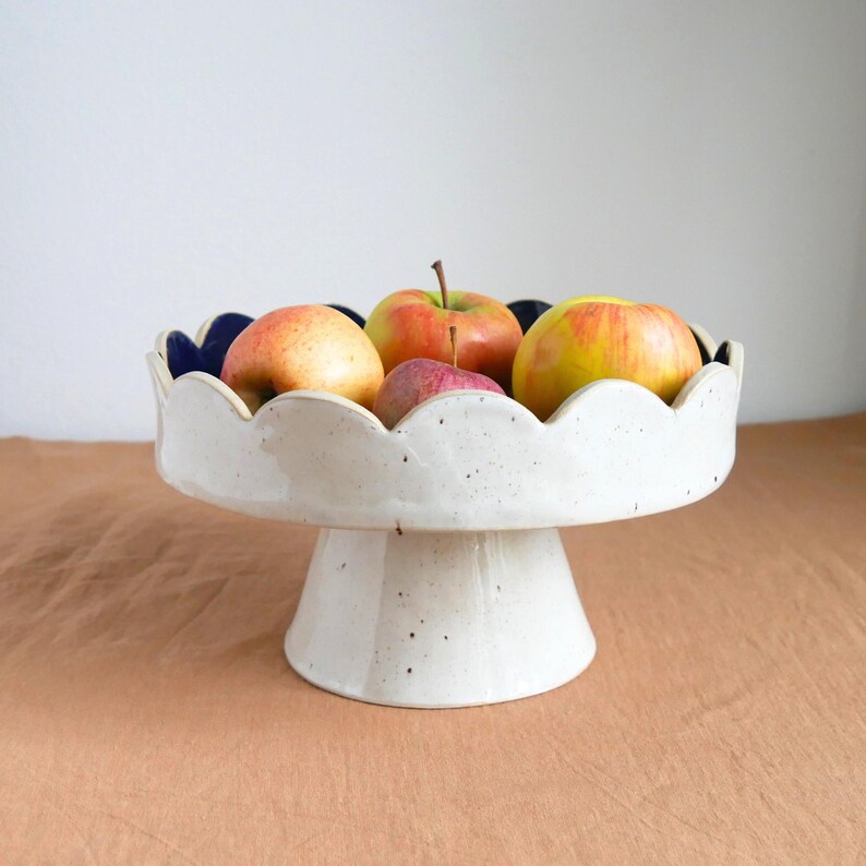 Op de afbeelding: Een witte keramische fruitschaal op een voet, met een geschulpte rand en een donkerblauwe binnenkant. De schaal bevat verschillende appels in rode, gele en groene tinten. De schaal staat op een bruin oppervlak.