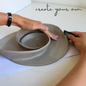 K&ouml;nnte beinhalten: Graues Keramikst&uuml;ck, das von Hand geformt wird. Das St&uuml;ck hat ein Spiraldesign mit einer zentralen Schale. Der Text "create your own" ist in Kursivschrift &uuml;ber dem Keramikst&uuml;ck geschrieben.