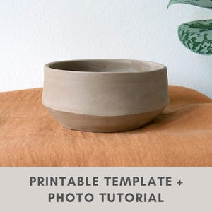 May include: A light brown ceramic bowl with a rounded shape and a slightly flared rim. The bowl is sitting on a textured orange fabric surface.