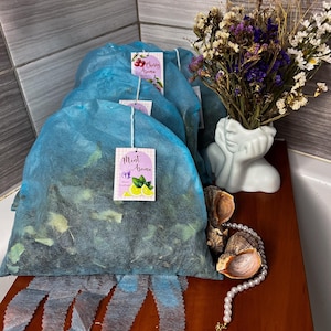 Puede incluir: Varios saquitos de color turquesa llenos de hierbas secas, etiquetados como "Mint Aroma" y "Cherry Aroma", están dispuestos sobre una superficie de madera. Un pequeño jarrón blanco contiene un ramo de flores secas. También hay un collar de perlas y conchas marinas.