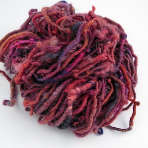 May include: A pile of hand-spun yarn in shades of deep red, pink, and purple. The yarn is made of natural fibers and has a rustic, hand-crafted look.