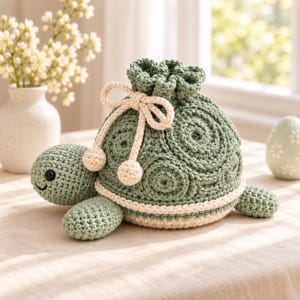 Può includere: Una pochette all'uncinetto a forma di tartaruga color verde salvia con chiusura a coulisse. Il guscio presenta un motivo circolare, mentre la testa, gli arti e la base sono realizzati in una tonalità di verde più chiara. Un cordino color crema con pompon chiude la parte superiore.