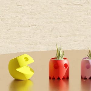 Puede incluir: Cinco macetas con forma de fantasma de colores diferentes con pequeñas plantas en su interior. Las macetas son rojas, rosas, azules, amarillas y verdes. Una figura amarilla de Pac-Man está en primer plano.