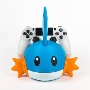 May include: A blue, white, and orange Mudkip-shaped controller holder with a white PlayStation controller in the background. The Mudkip has black eyes and orange fins. The controller has the standard PlayStation button layout.