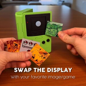 May include: A lime green handheld gaming device with a black screen and a selection of interchangeable display cards featuring Pok&eacute;mon characters. The text "SWAP THE DISPLAY" is visible.