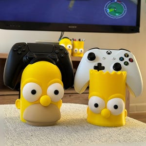 May include: Yellow Homer Simpson and Bart Simpson controller holders with black and white game controllers. The Homer holder has a black PlayStation controller, and the Bart holder has a white Xbox controller. The holders are on a white surface.