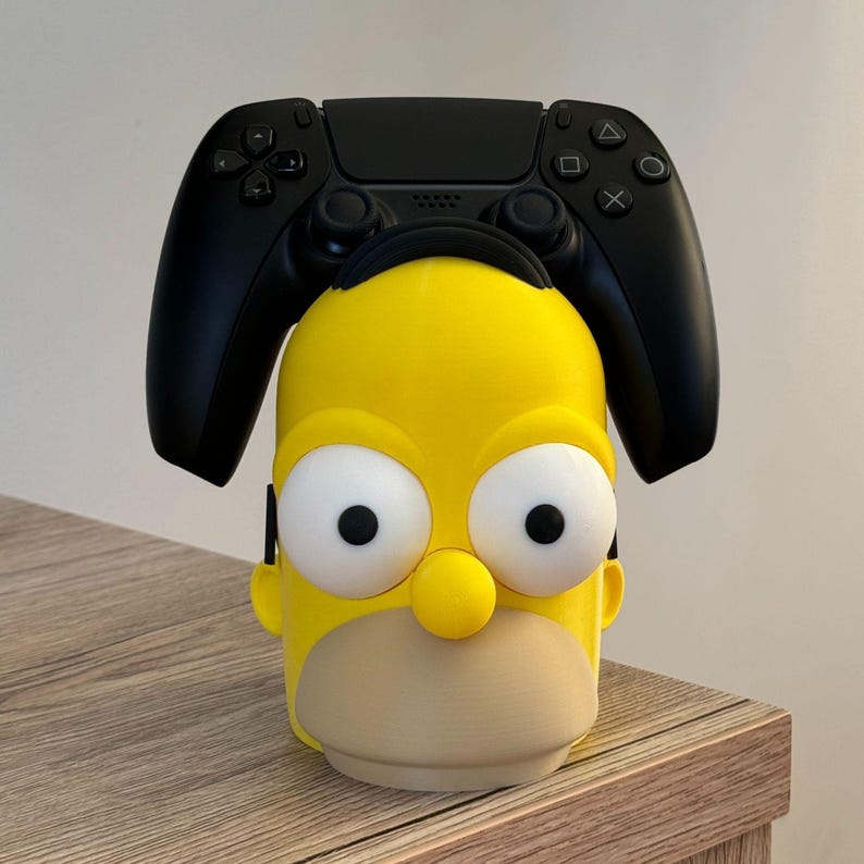 May include: A yellow and beige character-themed controller holder with a black controller resting on top. The holder is designed to resemble a cartoon character's head, with large white eyes and a prominent nose. The controller is black.