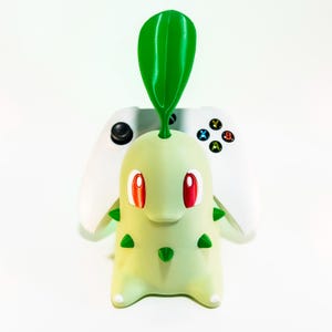 Può includere: Una statuetta di Chikorita verde chiaro con occhi rossi e punte verdi, con una grande foglia verde sulla testa. Un controller Xbox bianco è sullo sfondo.