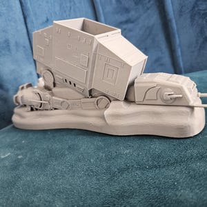 May include: A gray 3D printed model of a Star Wars AT-AT walker, with the top half of the walker tilted to the side. The model is sitting on a gray base.