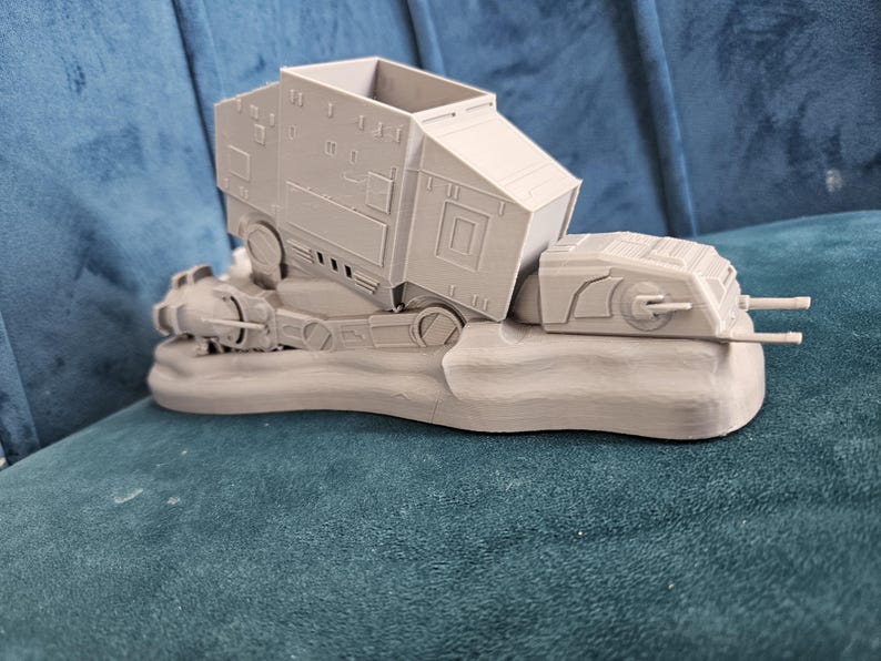 May include: A gray 3D printed model of a Star Wars AT-AT walker, with the top half of the walker tilted to the side. The model is sitting on a gray base.