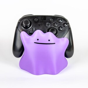 May include: A purple Ditto figure with a simple black facial expression is in front of a black Nintendo Switch Pro Controller. The controller has buttons and joysticks.
