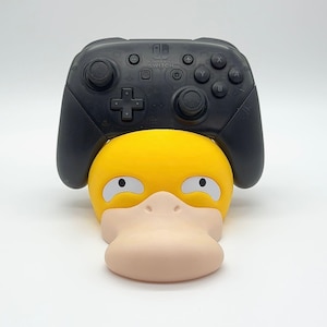 May include: A black Nintendo Switch Pro Controller rests on a yellow and tan Psyduck-shaped stand. The controller has buttons and joysticks. The stand has a duck-like face with white eyes and a tan beak.