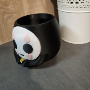May include: Black and white ceramic planter shaped like a cartoon character with pink cheeks and black eyes. The planter has a round opening at the top for planting.