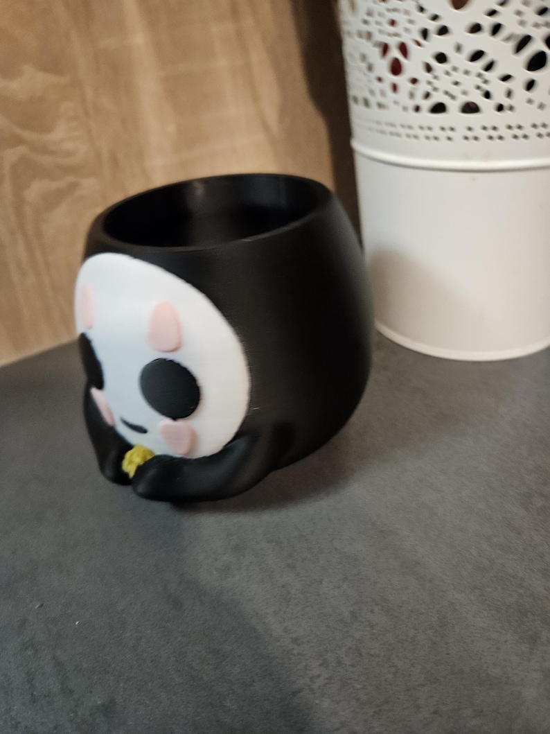 May include: Black and white ceramic planter shaped like a cartoon character with pink cheeks and black eyes. The planter has a round opening at the top for planting.