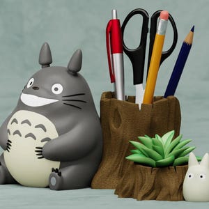 May include: A gray and white Totoro character figurine sits next to a brown wooden pencil holder with a green succulent plant. The pencil holder has a red pen, a pair of black scissors, a yellow pencil, and a blue pencil inside. A smaller white Totoro figurine sits in front of the pencil holder.