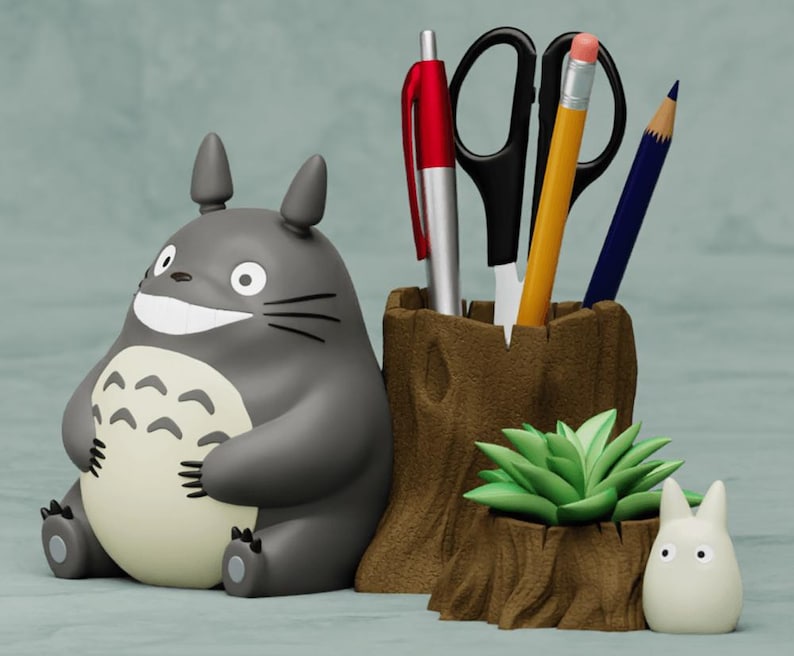 May include: A gray and white Totoro character figurine sits next to a brown wooden pencil holder with a green succulent plant. The pencil holder has a red pen, a pair of black scissors, a yellow pencil, and a blue pencil inside. A smaller white Totoro figurine sits in front of the pencil holder.