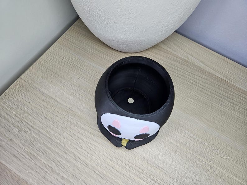 May include: Black and white planter shaped like a cartoon character with a round head and a small mouth. The planter has a small hole in the bottom for drainage.