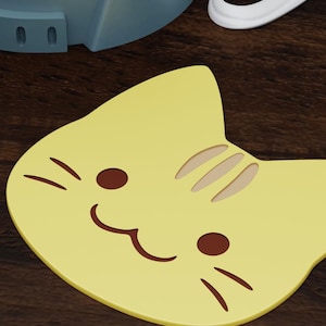 May include: A yellow cat-shaped coaster with a brown smiling mouth and brown whiskers. The coaster has a brown stripe pattern on the back.
