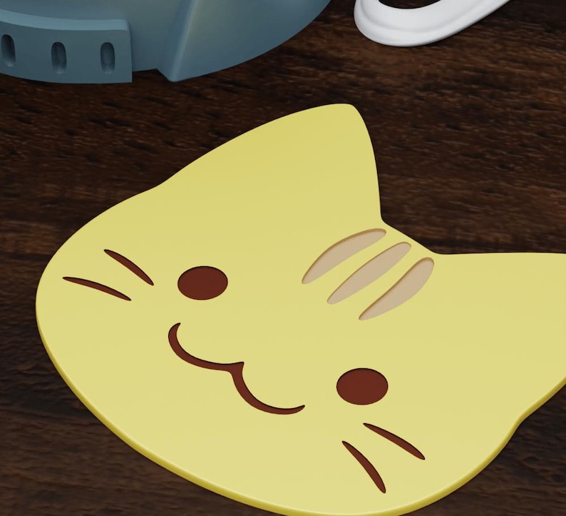 May include: A yellow cat-shaped coaster with a brown smiling mouth and brown whiskers. The coaster has a brown stripe pattern on the back.