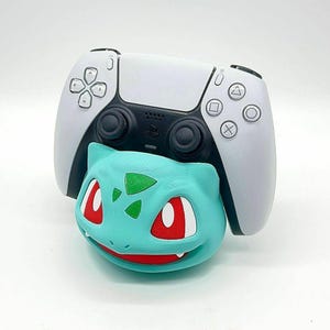 Puede incluir: Un mando blanco y negro de PlayStation 5 descansa sobre un soporte para mando con tem&aacute;tica de Bulbasaur, de color turquesa. El soporte de Bulbasaur presenta ojos rojos y un dise&ntilde;o de hoja verde. El mando tiene la distribuci&oacute;n de botones est&aacute;ndar.