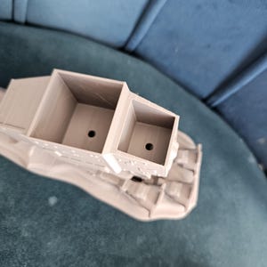May include: A 3D printed model of a vehicle with a gray finish. The vehicle has a large, open cargo bed with two compartments. The model is sitting on a blue surface.