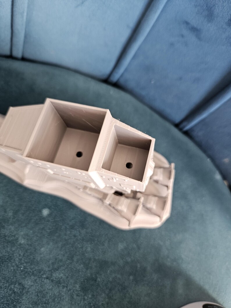 May include: A 3D printed model of a vehicle with a gray finish. The vehicle has a large, open cargo bed with two compartments. The model is sitting on a blue surface.