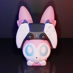 May include: A pink and white character-themed controller stand with a PlayStation 5 controller resting on top. The stand has large ears and a cute face. The controller is black and white.