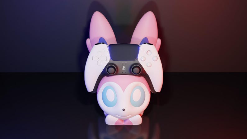 May include: A pink and white character-themed controller stand with a PlayStation 5 controller resting on top. The stand has large ears and a cute face. The controller is black and white.
