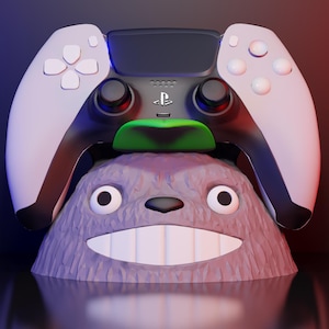 Pode incluir: Um controle PlayStation 5 repousa sobre um suporte roxo inspirado em Totoro. O controle é branco e preto com o logotipo da PlayStation. O suporte tem um rosto sorridente e olhos grandes. O fundo é um gradiente de azul e vermelho.