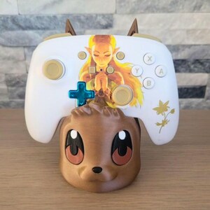May include: A white video game controller with gold buttons and a princess graphic, resting on a brown, cartoon-style character base. The controller has a blue directional pad and is designed for gaming.