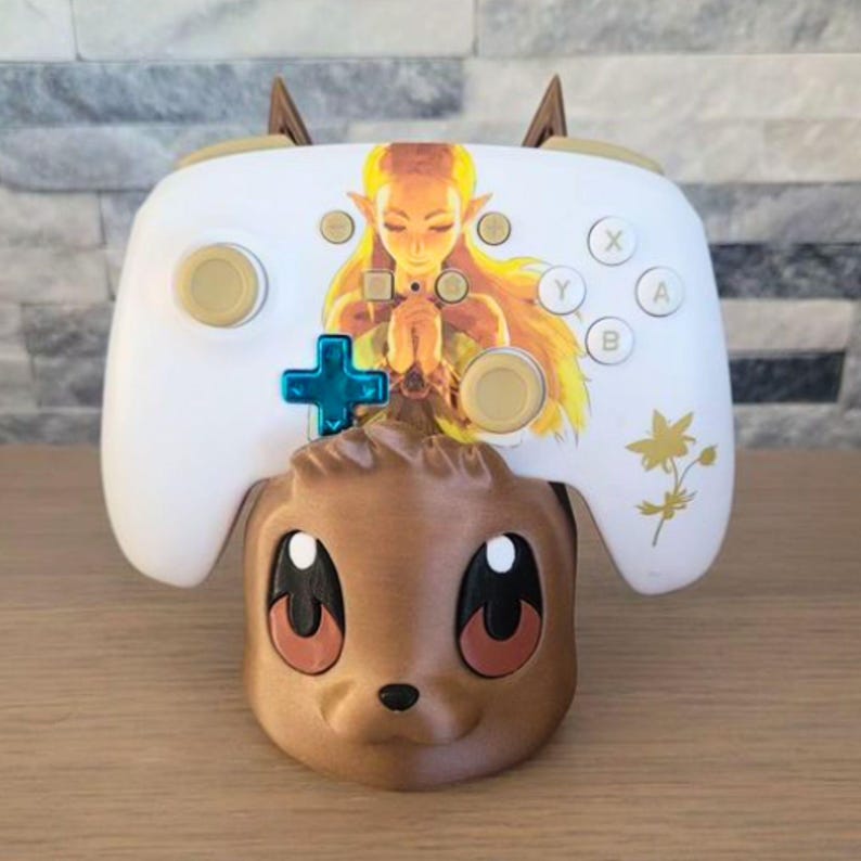 May include: A white video game controller with gold buttons and a princess graphic, resting on a brown, cartoon-style character base. The controller has a blue directional pad and is designed for gaming.
