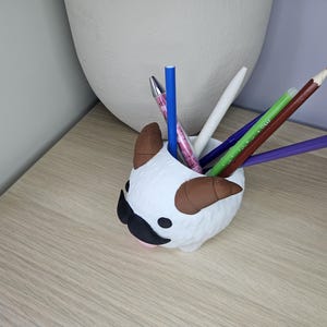 May include: White ceramic pencil holder shaped like a cartoon animal with brown horns and a black mustache. The holder is filled with colored pencils and pens.