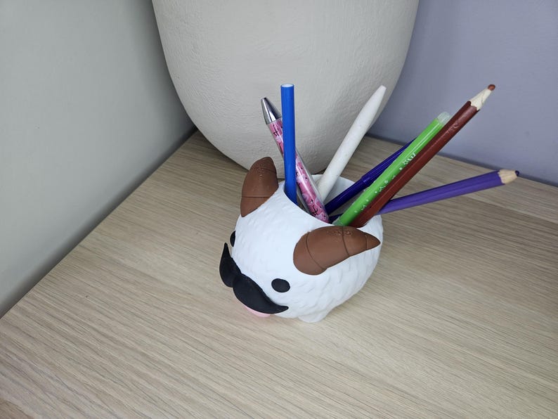 May include: White ceramic pencil holder shaped like a cartoon animal with brown horns and a black mustache. The holder is filled with colored pencils and pens.