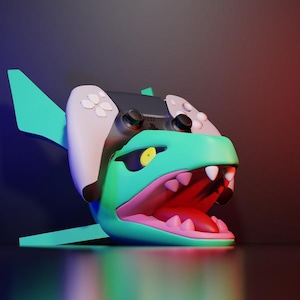 May include: A 3D rendering of a shark-shaped gaming controller. The shark is teal with a pink mouth and white teeth. The controller is black and white with buttons. The background is a gradient of blue and red.