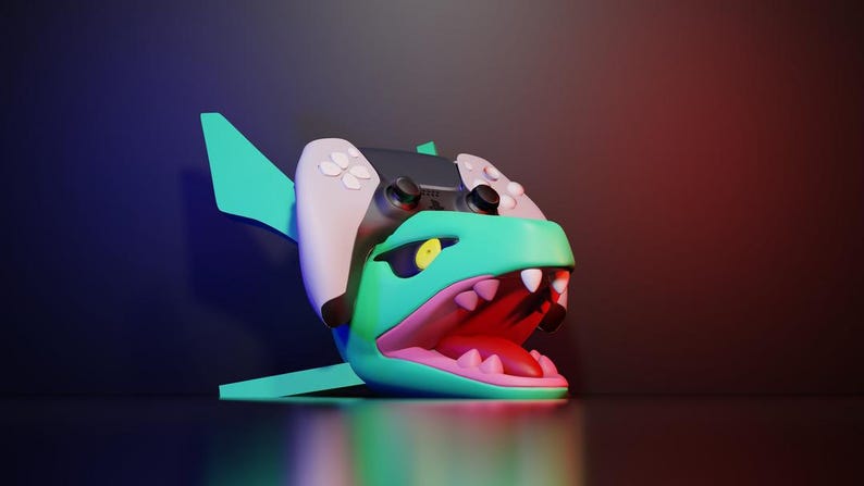 May include: A 3D rendering of a shark-shaped gaming controller. The shark is teal with a pink mouth and white teeth. The controller is black and white with buttons. The background is a gradient of blue and red.